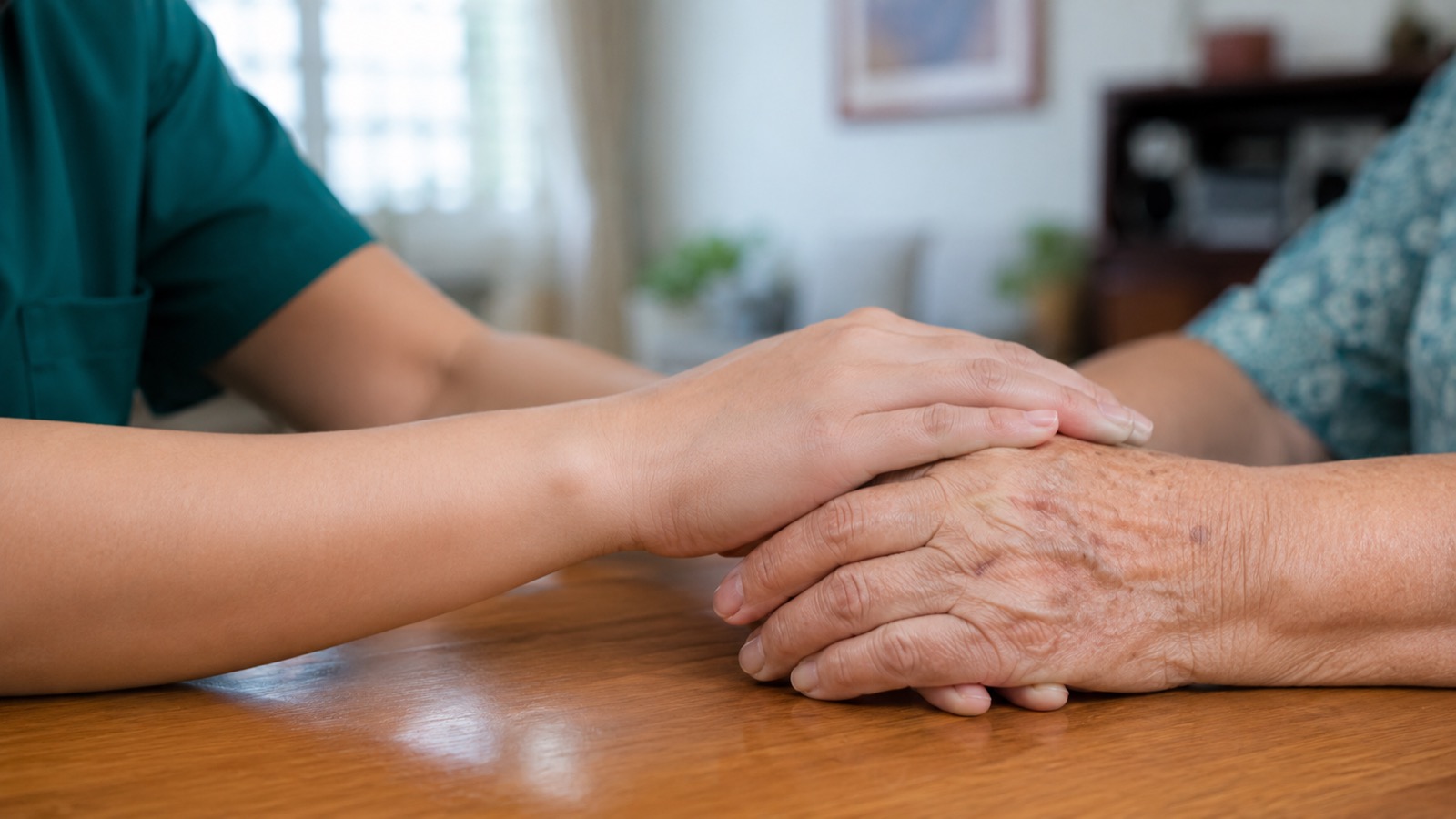 Close-up of a home nurse's hand resting reassuringly over an elderly patient's hands at a table at home in Singapore